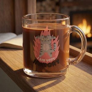 May include: A clear glass mug filled with a dark brown liquid, possibly coffee or hot chocolate. The mug features a cartoon cat design with the text "I Am very HUNGRY" in a red flame graphic. The mug has a large handle.