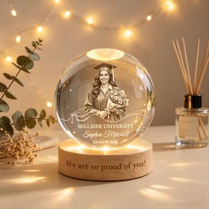 May include: A clear crystal sphere with a graduation portrait of a woman in a cap and gown, holding flowers. The sphere sits on a wooden base with the text "We are so proud of you!" and the name "Sophia Mitchell" and "Class of 2026".