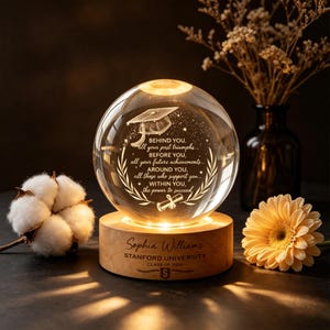 May include: A graduation-themed crystal ball with a wooden base, illuminated from within. The sphere is engraved with a graduation cap, text, and a laurel wreath. The base is engraved with a name, university, and class year. A flower and cotton ball are in the background.
