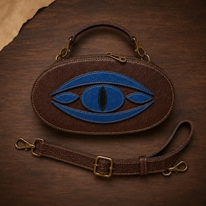Puede incluir: Un bolso ovalado de color marrón con una aplicación en forma de ojo azul y una pupila negra. El bolso incluye un asa superior y una correa de hombro marrón ajustable y extraíble, aseguradas con herrajes de metal en tono latón antiguo.