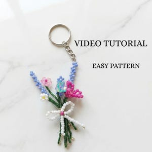 Può includere: Un portachiavi a bouquet di fiori di perline con un anello argentato. Il bouquet presenta fiori rosa, blu e bianchi di perline, steli verdi e un fiocco bianco. Il testo "VIDEO TUTORIAL" e "EASY PATTERN" sono visibili.