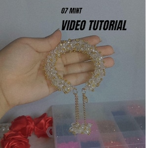 May include: A clear beaded bracelet with gold accents and a dangling charm is held in a hand. The bracelet is adorned with small, clear beads and gold-colored metal. The text "07 MINT VIDEO TUTORIAL" is in the upper left corner.