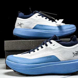 May include: Two athletic shoes, one on top of the other, against a black background. The shoes are white and blue, with the word "SYLAN" printed on the side. The shoes have a thick, light blue sole and a mesh upper.