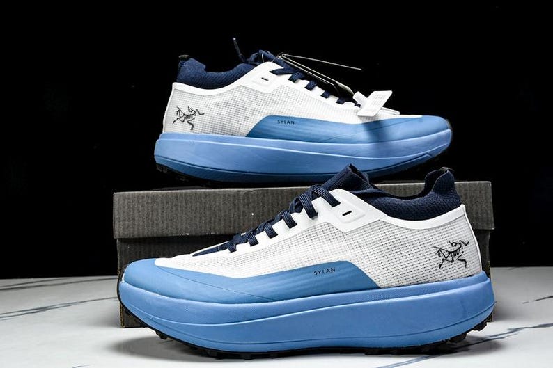 May include: Two athletic shoes, one on top of the other, against a black background. The shoes are white and blue, with the word "SYLAN" printed on the side. The shoes have a thick, light blue sole and a mesh upper.