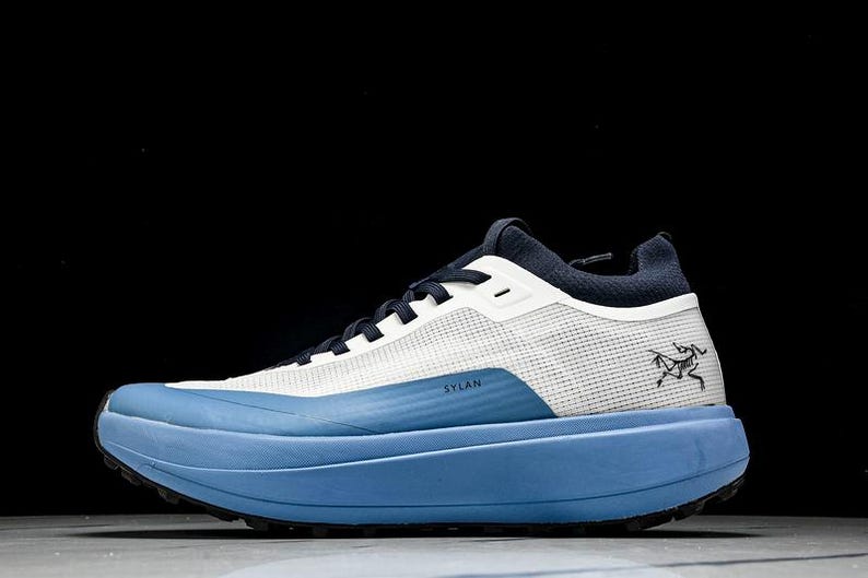 May include: A blue and white athletic shoe with the word "SYLAN" printed on the side. The shoe has a thick, light blue sole and a mesh upper. A black logo is on the side of the shoe.