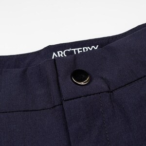 May include: Close-up of navy blue pants with a black button closure. The brand name "ARC'TERYX" is printed in white on the waistband. The fabric appears to be a smooth, durable material, suitable for outdoor activities.