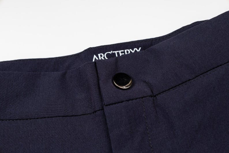 May include: Close-up of navy blue pants with a black button closure. The brand name "ARC'TERYX" is printed in white on the waistband. The fabric appears to be a smooth, durable material, suitable for outdoor activities.