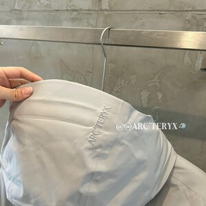 May include: A light gray hooded jacket with the brand name "ARC'TERYX" embroidered on the hood. The jacket is hanging on a metal hanger. The text "@@ARC'TERYX" is visible on the jacket.