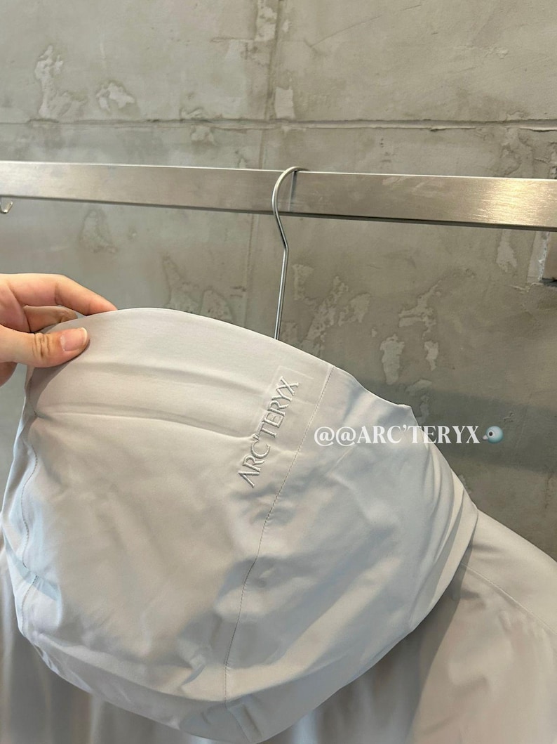May include: A light gray hooded jacket with the brand name "ARC'TERYX" embroidered on the hood. The jacket is hanging on a metal hanger. The text "@@ARC'TERYX" is visible on the jacket.