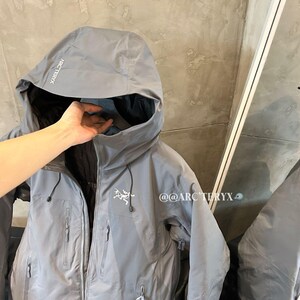 May include: A gray hooded jacket with multiple zippered pockets and the brand name "Arc'teryx" printed on the hood. The jacket has a Gore-Tex label and a small logo on the chest. The jacket is shown with a hand inside the hood.