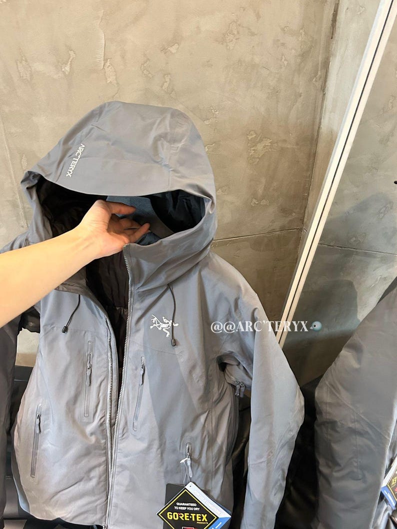 May include: A gray hooded jacket with multiple zippered pockets and the brand name "Arc'teryx" printed on the hood. The jacket has a Gore-Tex label and a small logo on the chest. The jacket is shown with a hand inside the hood.