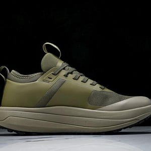 May include: Olive green athletic shoe with a thick, layered sole. The shoe features a mesh toe box and a pull tab on the heel. The laces and upper are a solid olive green color, with a neutral-toned sole.