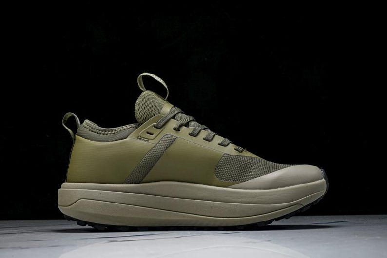 May include: Olive green athletic shoe with a thick, layered sole. The shoe features a mesh toe box and a pull tab on the heel. The laces and upper are a solid olive green color, with a neutral-toned sole.