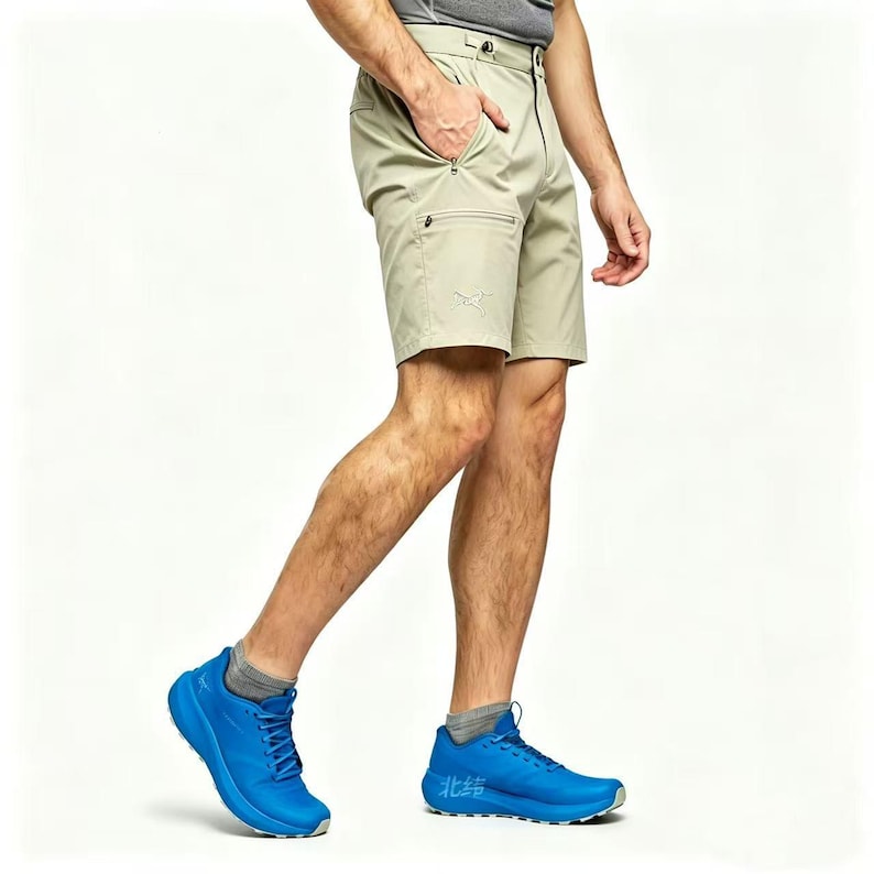 May include: Khaki-colored shorts with multiple pockets and a small embroidered logo. The shorts are paired with blue athletic shoes. The person is wearing a gray shirt and socks. The shorts have a zipper closure and a button.