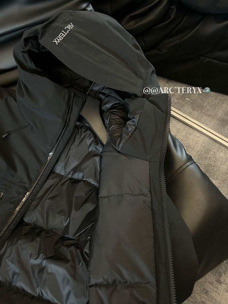 May include: Black hooded jacket with the brand name "Arc'teryx" printed on the hood. The jacket has a full zipper and a quilted interior. The jacket is made of a smooth, matte material.