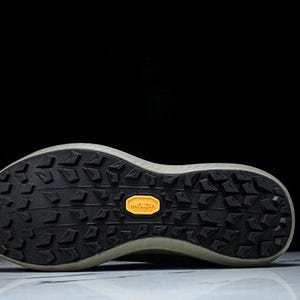 May include: Close-up of the sole of a running shoe. The sole is black with a textured pattern and a yellow Vibram logo. The shoe has a light-colored midsole and is set against a black background.