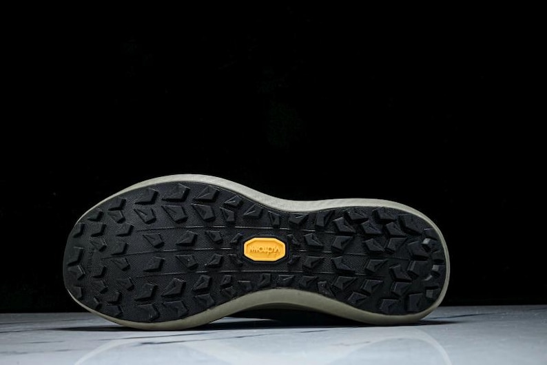 May include: Close-up of the sole of a running shoe. The sole is black with a textured pattern and a yellow Vibram logo. The shoe has a light-colored midsole and is set against a black background.