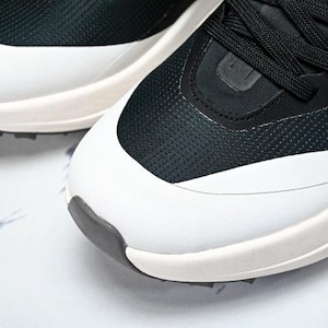 May include: Close-up of a pair of athletic shoes. The shoes feature a black mesh upper with white accents and a white sole. Black laces are visible. The toe and heel have a dark gray rubber detail. The shoes are on a white surface.