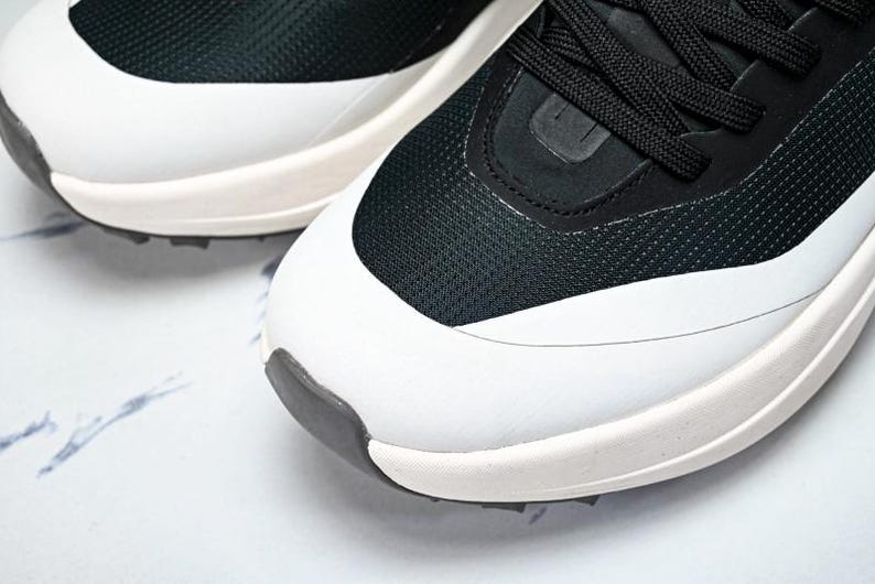 May include: Close-up of a pair of athletic shoes. The shoes feature a black mesh upper with white accents and a white sole. Black laces are visible. The toe and heel have a dark gray rubber detail. The shoes are on a white surface.