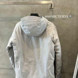 May include: A light gray hooded jacket hanging on a metal rack. The jacket has a full zipper and a hood. The brand name "ARC'TERYX" is visible on the hood. The background is a light gray wall.