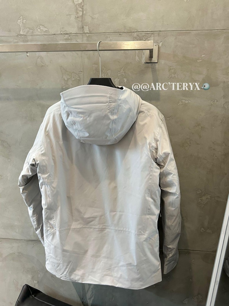 May include: A light gray hooded jacket hanging on a metal rack. The jacket has a full zipper and a hood. The brand name "ARC'TERYX" is visible on the hood. The background is a light gray wall.