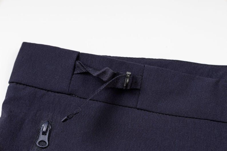 May include: Close-up of navy blue athletic pants. The image highlights the waistband with a drawstring, a metal clasp, and a zipper pocket. The fabric appears to be a textured, technical material.