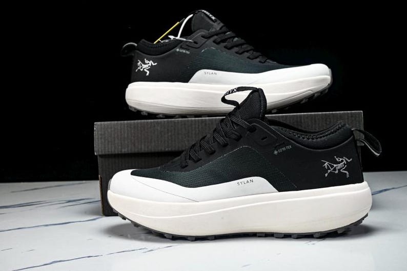 May include: Black and white athletic shoes with thick soles. The shoes have a black upper with white accents and a white sole. The brand logo is visible on the side. The word "SYLAN" is printed on the side of the shoe.