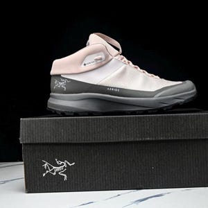 May include: A light pink and gray hiking shoe with the word "AERIOS" printed on the side, resting on a black box. The shoe has a mesh upper and a gray sole. The box has a white logo on the front.