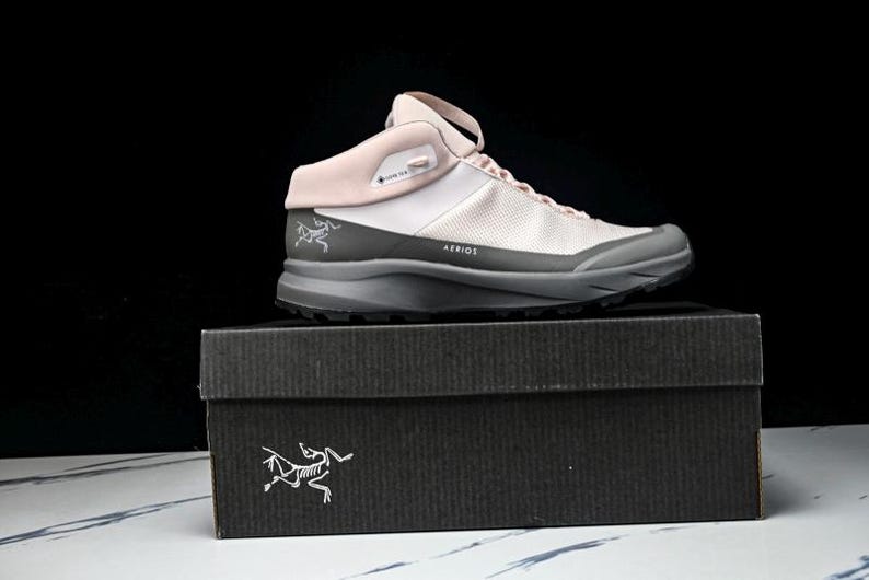 May include: A light pink and gray hiking shoe with the word "AERIOS" printed on the side, resting on a black box. The shoe has a mesh upper and a gray sole. The box has a white logo on the front.