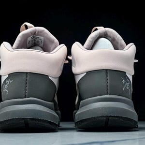 May include: Pair of gray and white athletic shoes with a light pink collar and tongue. The shoes have a thick gray sole and a small logo on the heel. The background is black.