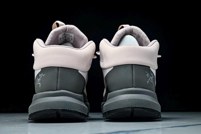 May include: Pair of gray and white athletic shoes with a light pink collar and tongue. The shoes have a thick gray sole and a small logo on the heel. The background is black.