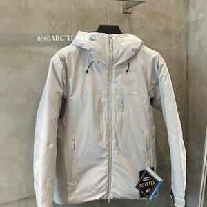 May include: A light gray hooded jacket with a full-length zipper, multiple zippered pockets, and the Gore-Tex logo. The jacket is hanging on a hanger against a gray wall. The brand name is visible on the jacket.