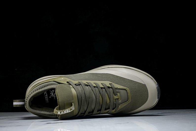 May include: Olive green athletic shoe with a mesh upper and a thick sole. The shoe features a lace-up closure and the brand name "Arc'teryx" is visible on the side. The shoe is designed for outdoor activities.