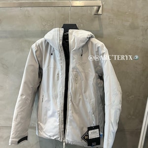 May include: A light gray hooded jacket with a full-length zipper, multiple pockets, and a black interior. The jacket is hanging on a metal rod against a gray wall. The brand name is visible.