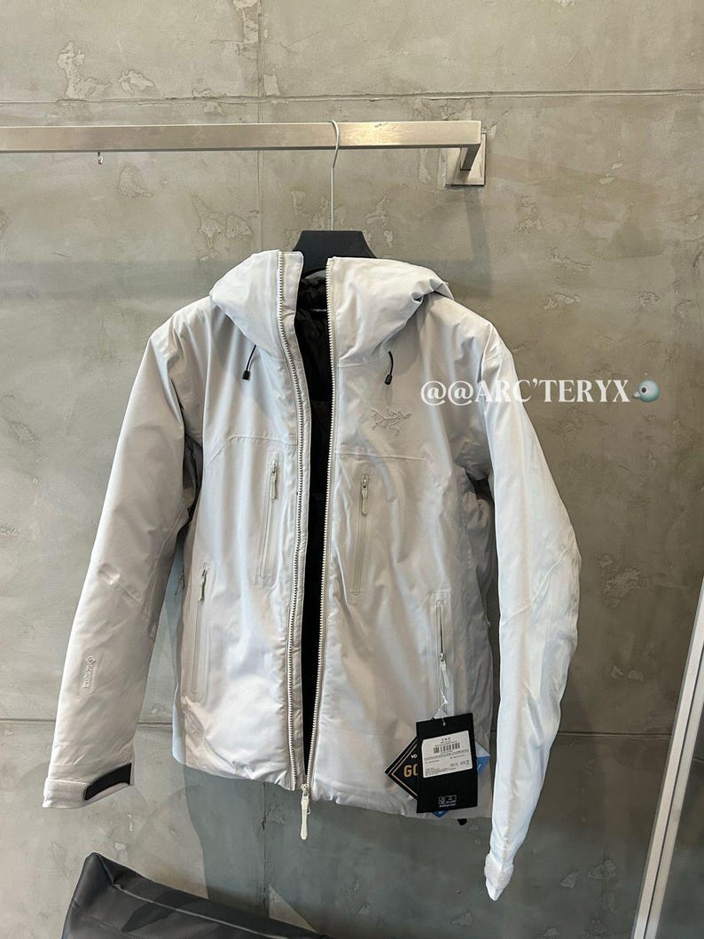 May include: A light gray hooded jacket with a full-length zipper, multiple pockets, and a black interior. The jacket is hanging on a metal rod against a gray wall. The brand name is visible.
