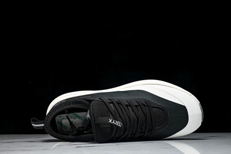 May include: Black and white athletic shoe with the word "TERYX" on the side. The shoe has a black upper with white accents and a white sole. The shoe is lying on its side against a black background.
