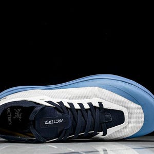 May include: A blue and white athletic shoe with a mesh upper and navy accents. The shoe features a blue toe cap and a navy tongue with the brand name "Arc'teryx" visible. The laces are navy, and the sole is white and blue.