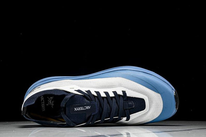 May include: A blue and white athletic shoe with a mesh upper and navy accents. The shoe features a blue toe cap and a navy tongue with the brand name "Arc'teryx" visible. The laces are navy, and the sole is white and blue.