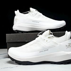 Puede incluir: Un par de zapatillas deportivas blancas con suelas negras, expuestas sobre una caja. Las zapatillas tienen el texto "VERTEX ALPINE" y un logotipo. Zapatillas dise&ntilde;adas para actividades al aire libre.