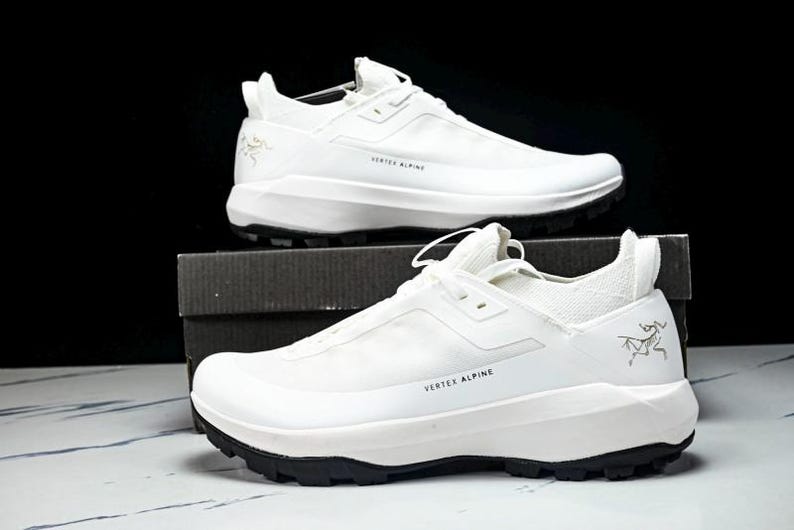 Puede incluir: Un par de zapatillas deportivas blancas con suelas negras, expuestas sobre una caja. Las zapatillas tienen el texto "VERTEX ALPINE" y un logotipo. Zapatillas dise&ntilde;adas para actividades al aire libre.