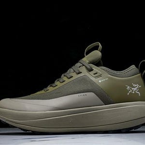 May include: Olive green athletic shoe with a thick sole. The shoe features a mesh upper, a pull loop, and the word "SYLAN" printed on the side. A white logo is visible on the side of the shoe. The shoe is designed for outdoor activities.