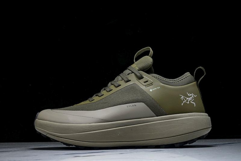 May include: Olive green athletic shoe with a thick sole. The shoe features a mesh upper, a pull loop, and the word "SYLAN" printed on the side. A white logo is visible on the side of the shoe. The shoe is designed for outdoor activities.