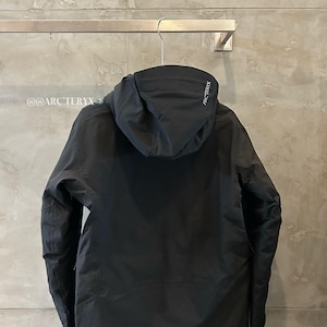 May include: Black hooded jacket hanging on a metal rack. The jacket has a minimalist design with a hood and long sleeves. The brand name "ARC'TERYX" is visible on the hood. The text "@@ARC'TERYX" is visible on the left side.