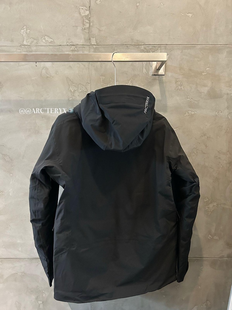 May include: Black hooded jacket hanging on a metal rack. The jacket has a minimalist design with a hood and long sleeves. The brand name "ARC'TERYX" is visible on the hood. The text "@@ARC'TERYX" is visible on the left side.