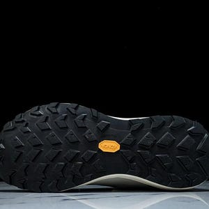May include: Close-up of the sole of a white and black athletic shoe. The black sole features a textured pattern with a small orange Vibram logo. The shoe is designed for traction and durability.