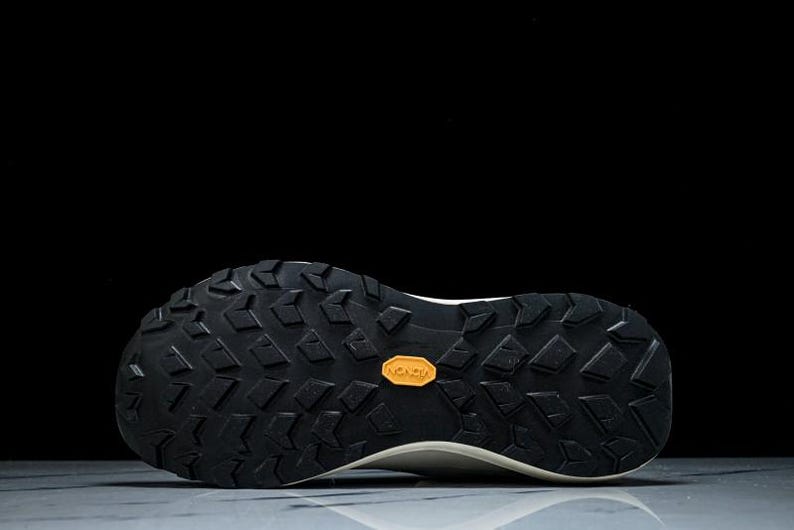 May include: Close-up of the sole of a white and black athletic shoe. The black sole features a textured pattern with a small orange Vibram logo. The shoe is designed for traction and durability.