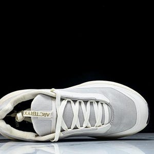 May include: A light gray athletic shoe with white laces and a white sole. The shoe has the brand name "Arc'teryx" printed on the tongue. The shoe is designed for athletic activities and has a modern, streamlined look.
