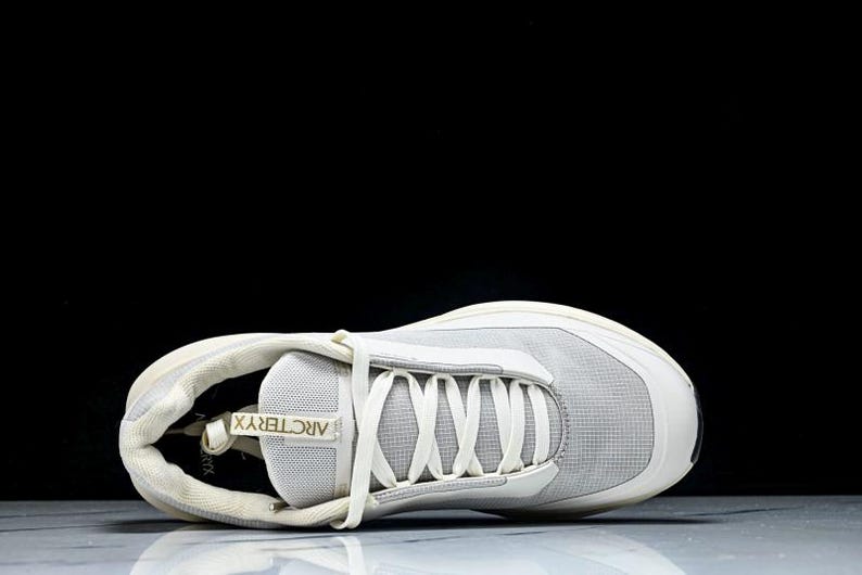 May include: A light gray athletic shoe with white laces and a white sole. The shoe has the brand name "Arc'teryx" printed on the tongue. The shoe is designed for athletic activities and has a modern, streamlined look.