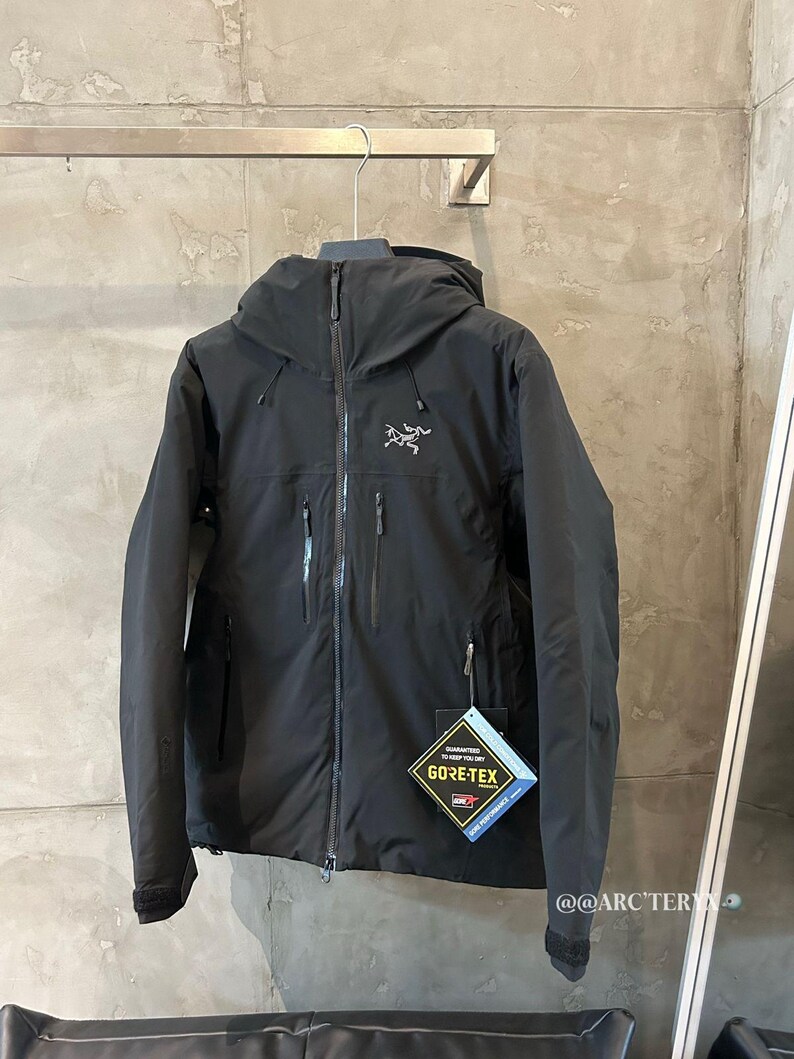 May include: Black hooded jacket with a zipper closure and multiple pockets. The jacket features the Gore-Tex logo and a small logo on the chest. The jacket is hanging on a metal hanger.