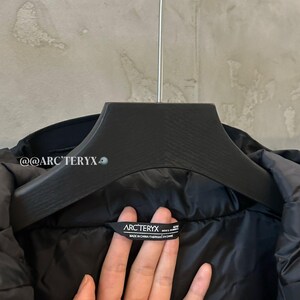 May include: Black jacket hanging on a black hanger. The jacket has a label that reads "ARC'TERYX" and "MADE IN CHINA". The jacket is made of a smooth, shiny material.